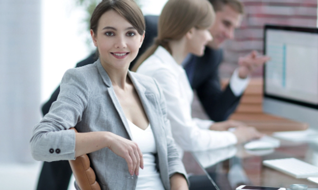 A woman with her elbow on a chair and two people blurred out in the background talking about something they are looking at on their computer