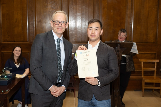 man in suit wearing tie and glasses standing beside student wearing a blazer and holding a certificate
