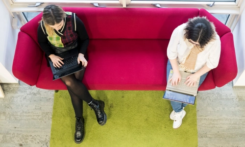 Two students sitting on a couch, working on laptops.