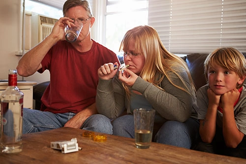 Two adults and one child sitting beside each other. One adult is drinking, the adult is smoking. The child is watching tv