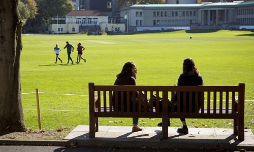 Students sitting on a bench by the cricket pitch in Trinity College