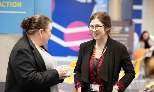 Two women talking at an event. The woman on the left is holding a ceramic tea-cup and saucer.