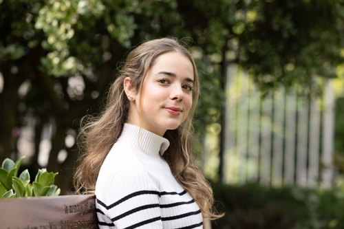 Irene Klotz Maroto sitting on a bench. Photo is taken to her right, and she is looking at the camera. Trees and green foliage blurred in background.