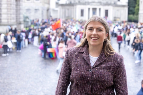 Photo of Jenny Salmon standing on the steps of a building in Trinity Front Square, smiling at the camera. Crowd of people blurred out in the background.