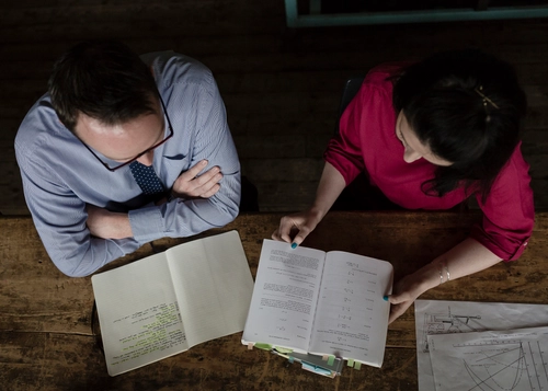 Two students sit at a wooden table reviewing documents, with one open notebook and one open book marked with tab