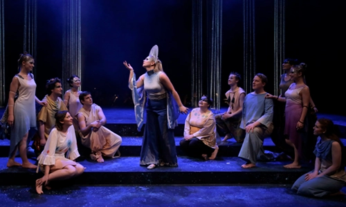 An image from sa stage production. We can see a group of people sitting on the stage, with in the middle a woman with a crown and toga around her talking.