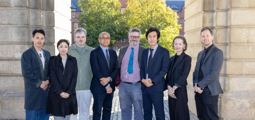 Eight participants pose for a group photograph beneath the historic stone archway at Trinity College Dublin. Professor Nathan Hill stands near the centre. Sunlit trees, cobblestones and a red-brick university building form the background.