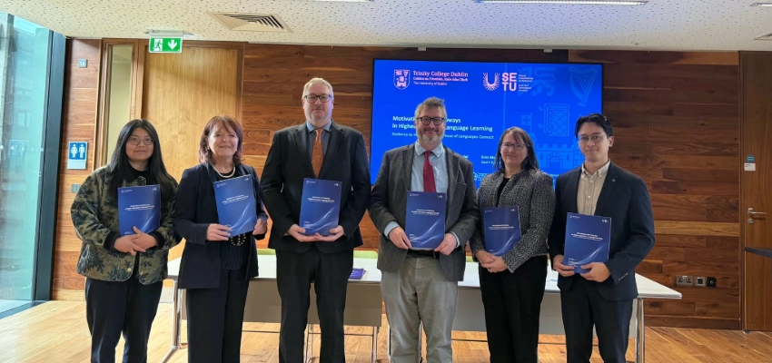Hongfei Wang, Karen Ruddock (Post-Primary Languages Ireland), Mark Bohan (Department of Education and Youth), Nathan Hill, Erin McNamara Cullen and Rowland Imperial hold copies of the Motivations and Pathways in Higher Education Language Learning research report at Trinity College Dublin, standing before a presentation screen displaying the report title.