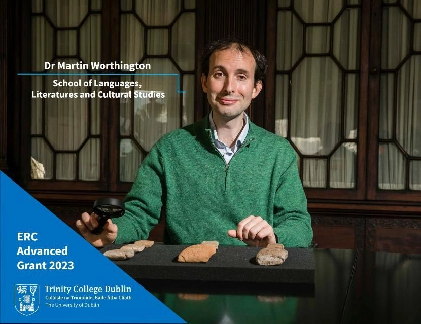 A man in a green jumper in front of a decorative bookcase, with stone artefacts set out in front of him