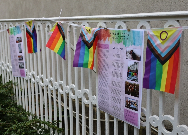 Image of pride flag bunting along a white railing. Pages are pinned between the flags that detail the 'Pride at Trinity' timeline from 1980 to 2022.