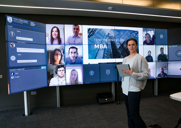 A person standing in front of a wall sized large monitor conducting an online conference