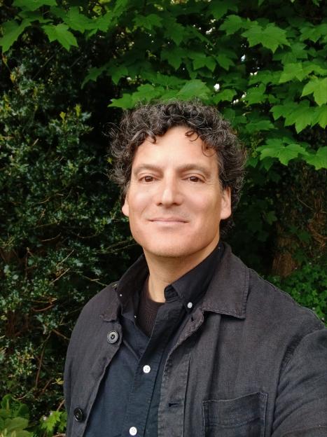 Male figure standing outdoors, head and shoulders, green plants in the background.