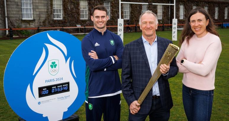 Three people stand facing the camera with a countdown clock marking 100 days until the Olympic and Paralympic Games. one of the people is holding an Olympic torch