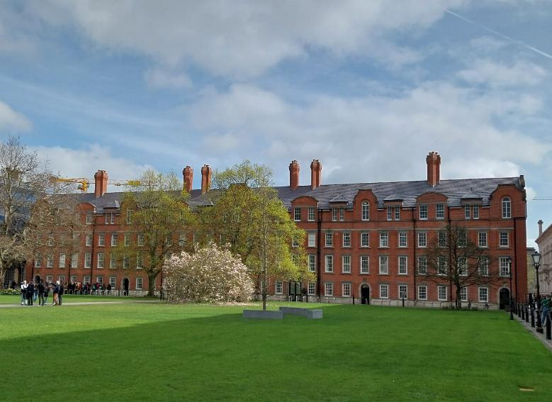 Trinity's red-brick Rubrics with the dark Valentia slate on their roofs. The Rubrics stand against a lovely blue sky, with green grass and beautiful trees on the campus lawns.