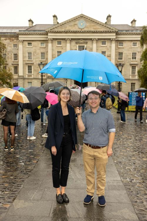 Drs Johanna Vos and Michael-John Dolan stand under a bright blue Trinity College Dublin umbrella in a rainy Front Square.
