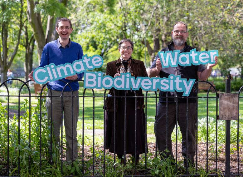 Prof Ed Hawkins, Prof Yvonne Buckley and Prof Mark Emerson holding light blue signs reading Climate, Biodiversity and Water. They are standing against a black railing, with beautiful trees in the background and sun pouring through.