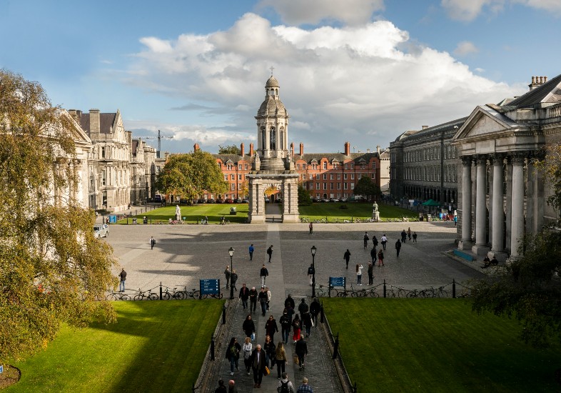 Front Square at Trinity College Dublin featuring the iconic Campanile, surrounding historic buildings, open courtyard, and people walking across the square, under a blue sky