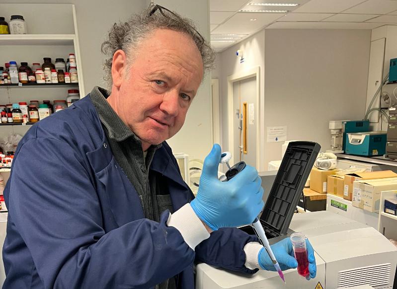 Prof. Gavin Davey wearing a blue lab coat and gloves works with a pipette in the laboratory.