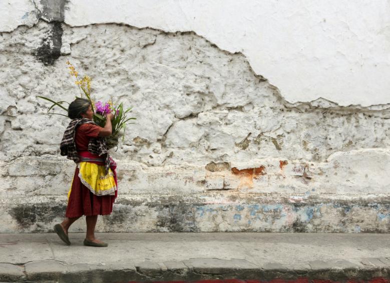 A Guatemalan woman walks in colourful clothes past a stone wall, carrying colourful flowers.