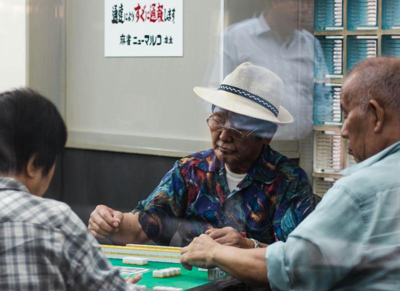 Three elderly Japanese men sit at a table playing a board game. A sign in Japanese is on the wall behind.