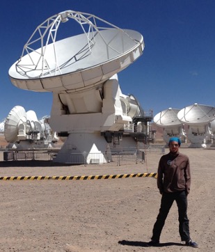 Prof. Luca Matra wearing dark clothes, standing on desert sand with a huge white telescope radar behind him, against a bright blue sky.