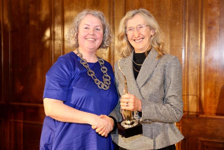 Provost Linda Doyle in a blue dress gives the Provost Innovation Award to Professor Orla Hardiman, who is wearing a black and white dress.