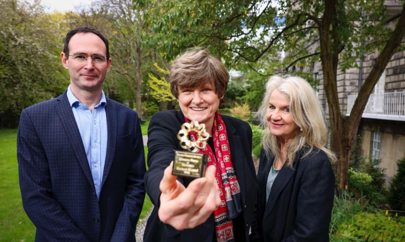 Prof Matthew Campbell, Dr Katalin Kariko with her gold DNA Dawson prize, and Prof Jane Farrar in Trinity with beautiful trees and plants in the background.