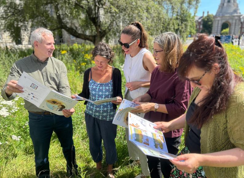 Prof Tom Curran, Prof Catherine Farrell, Dr Máire Nic an Bhaird, Prof Jane Stout and Dr Laoise Ní Chléirigh reading the comic in Trinity's Front Square at the launch of the comic, with bright blue sky and green foliage behind them.