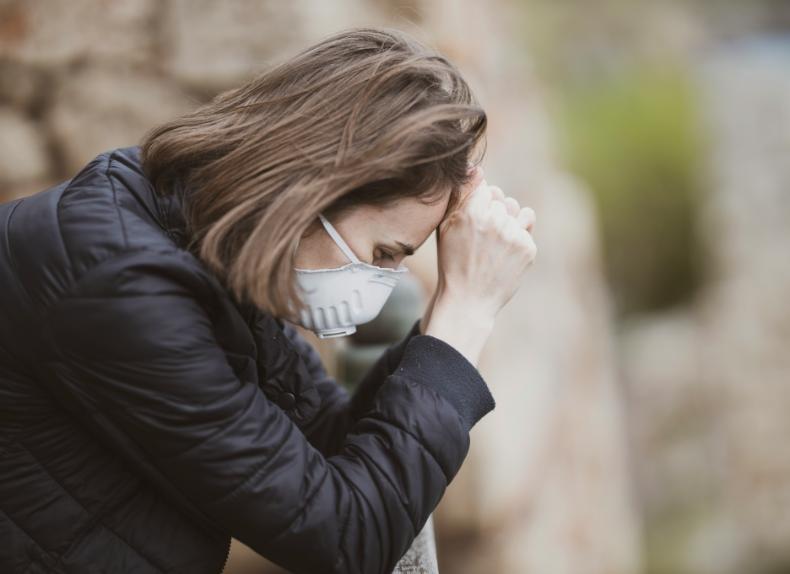 A dark-haired woman wearing a black jacket and a face mask coughs while sitting on a bench.