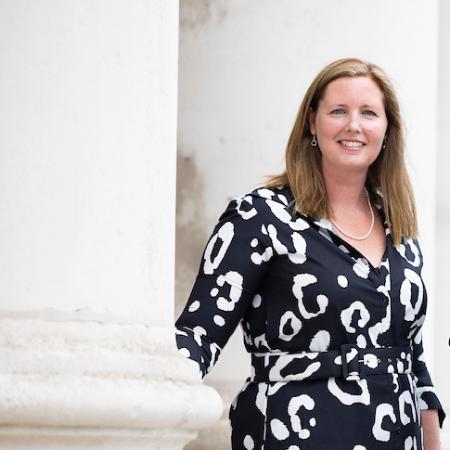 Dr Sarah Doyle in a black and white dress standing on the steps of the Exam Hall in Trinity