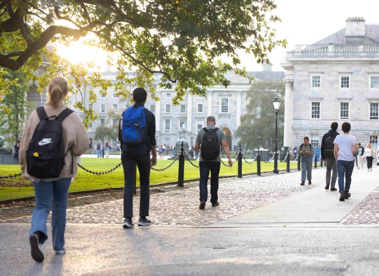 Students walk towards Front Gate in the distance on a sunny day in Trinity College Dublin. Sunlight is streaming through a gap in the branches of a tree, with the campanile and Front Square in the background.