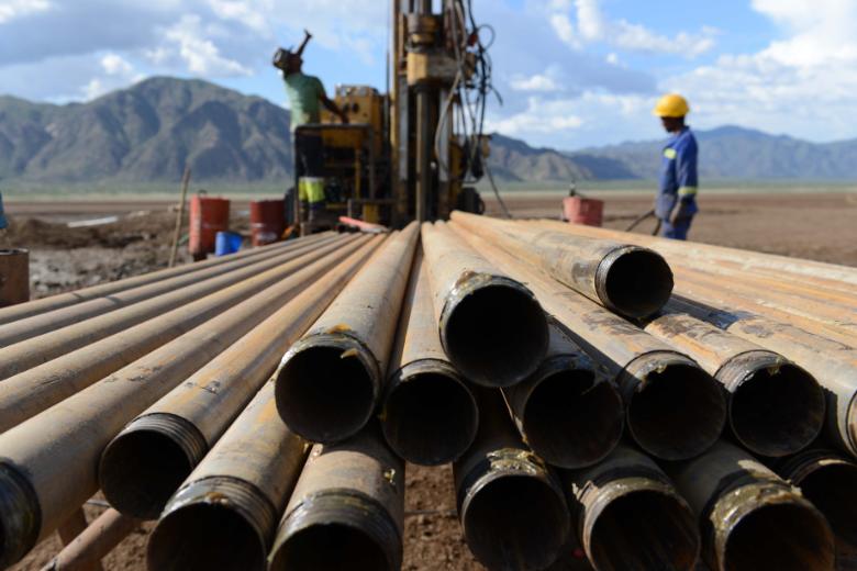 Pipes used for drilling in Chew Bahir. A worker wearing a hard hat is in the background with other machinery and mountainous peaks in the distance.
