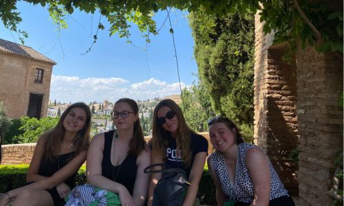 Four young woman sit in a garden surrounded by greenery with a blue sky behind them
