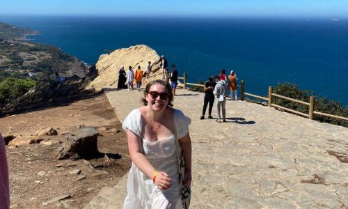 A young woman wearing sunglasses and a white dress, behind her is a blue sky and sea