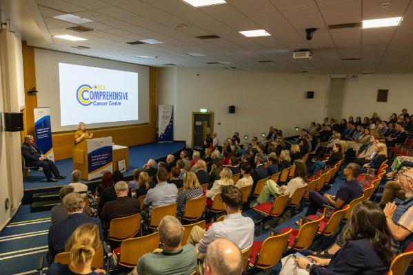 Linda speaking to a full lecture theatre. There is a slide projected onto the screen behind her which reads Comprehensive Cancer Centre.