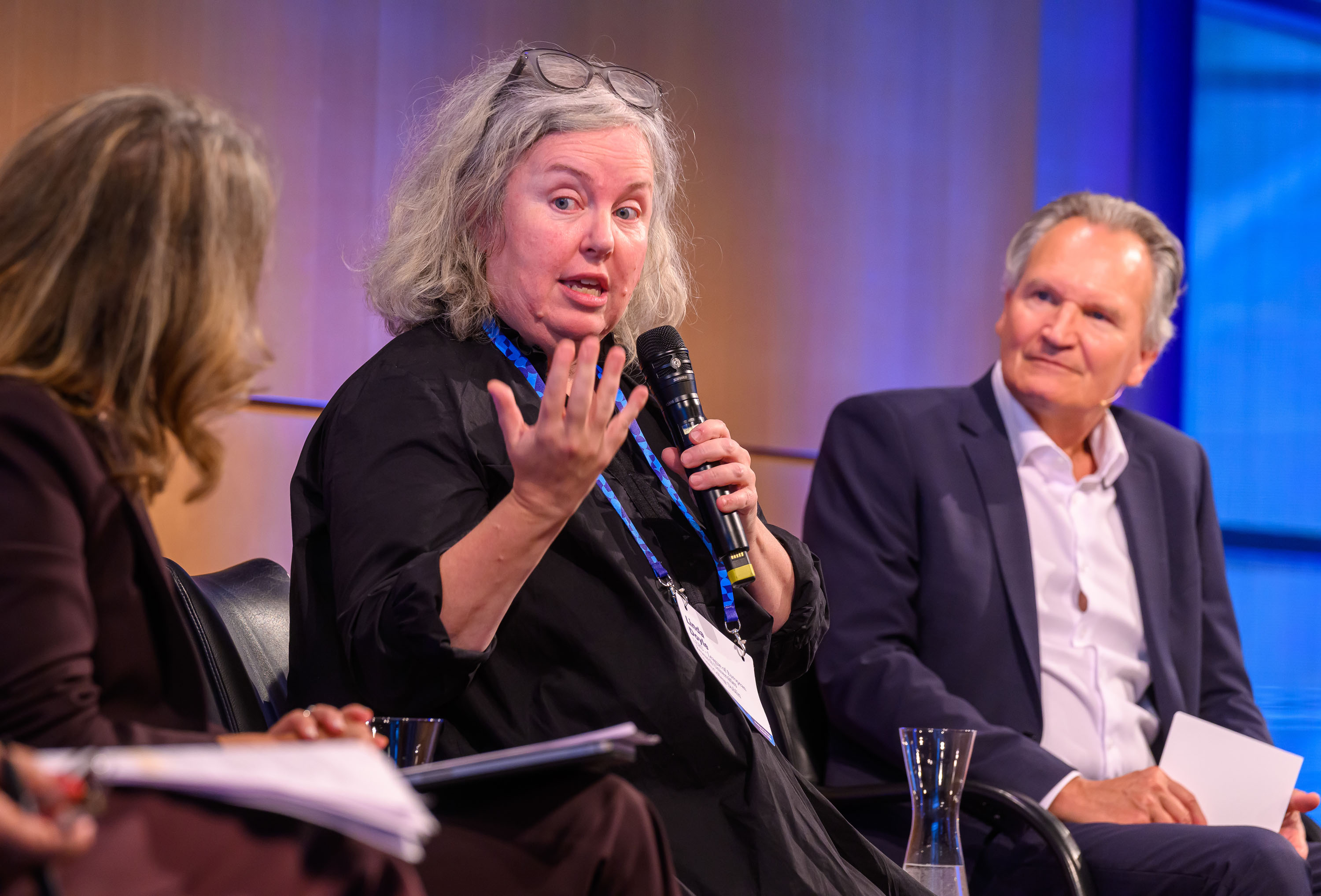 Close-up of Linda speaking on stage at the Hamburg Science Summit. She is speaking into a mic and gesturing towards the panelist to her right, who is mostly out of frame. The panel MC is visible, seated to her left.