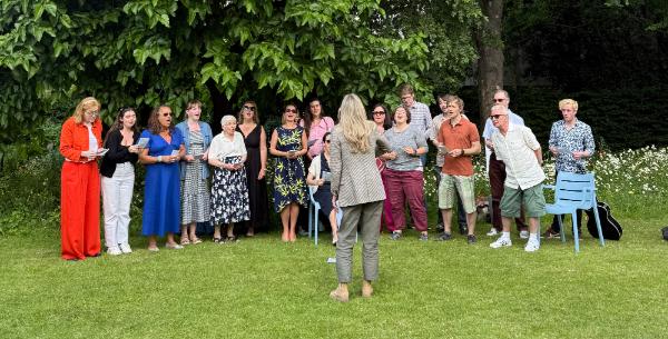 The AHSS Choir are mid-performance, standing in a line on the lawn of the Provosts garden. Their conductor stands in front of them. Trees in the background.