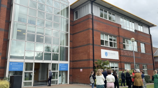The outside of TSJCI with people standing on a lawn to the right of the frame. The 3-storey red-brick building has the Trinity logo on its facade, and a tall, conservatory-style entrance way that runs up the entire height of the building.