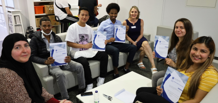 Five learners holding up their completion certificates in the Global Room, sitting with two Trinity volunteers.