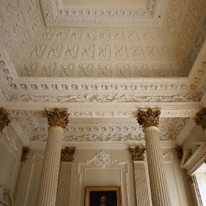 Photo taken looking up at the ceiling of the Provost's Saloon. The walls and ceiling are cream, and are covered in ornate plasterwork. There are four cream columns with gold detail at the top, and a portrait hanging in the middle.