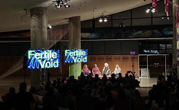 Photo taken from the back of a dark, large room. There is a stage at the front of the room with 4 women sitting under lights. They are holding mics and talking to each other. There is a seated audience watching them, and two TV screens to the left which read 'Fertile Void'.