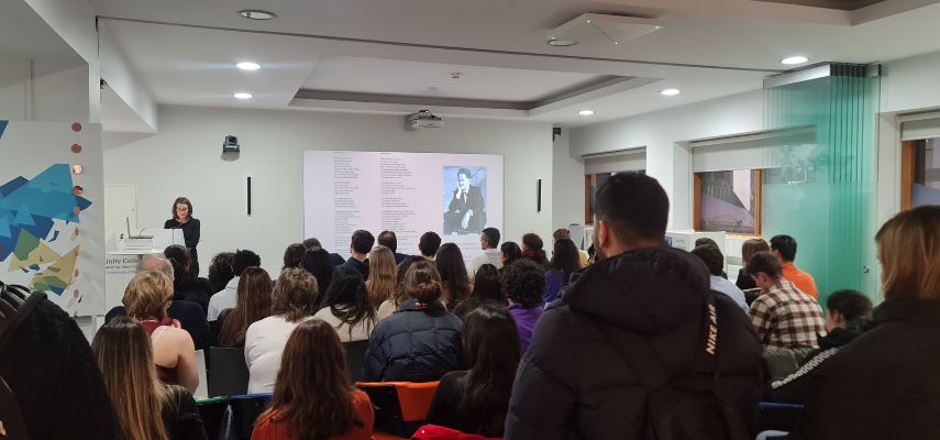 A speaker stands at a podium in at the front of the room, with a slide show on their right