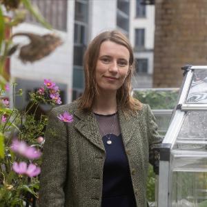 Photo of Eleanor Mullen standing in an urban garden. There is a green house to her right, and pink flowers to her left.
