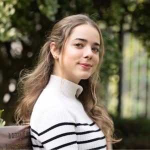 Irene Klotz Maroto sitting on a bench. Photo is taken to her right, and she is looking at the camera. Trees and green foliage blurred in background.