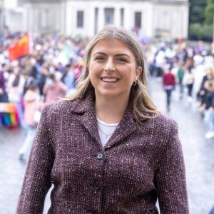 Photo of Jenny Salmon standing on the steps of a building in Trinity Front Square, smiling at the camera. Crowd of people blurred out in the background.