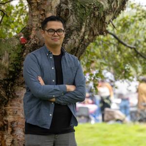 Photo of Nathan Cruz standing in front of a tree. He has his arms folder and is looking at the camera.
