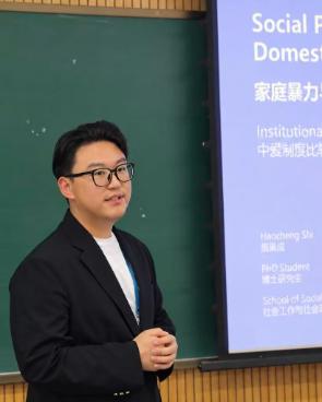 A man wearing a black blazer with glasses and black hair standing in front of a blackboard and screen