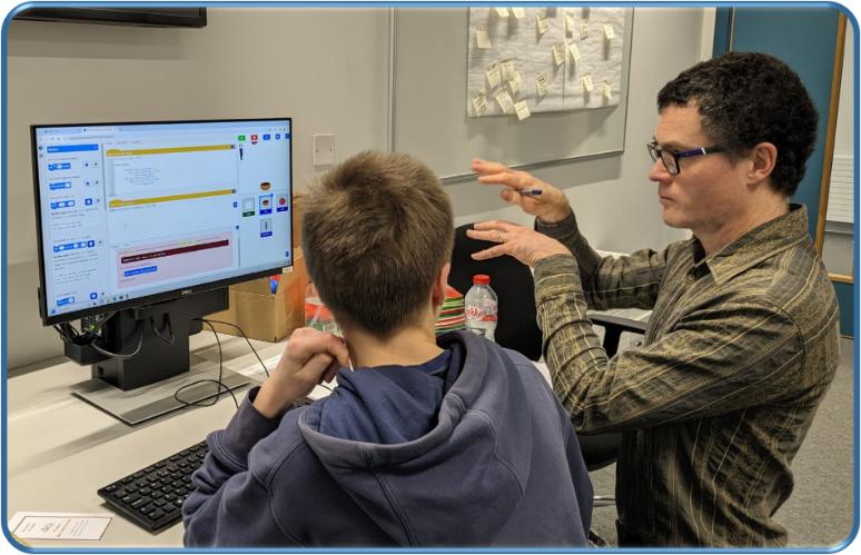 Dr Ben North in a muted green hoodie, talking to a student working in the Pytch app, on a desktop computer