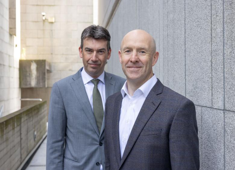 Dragos Tudorache, MEP, and Prof. John D Kelleher in suits and ties, standing against a grey brick backdrop.
