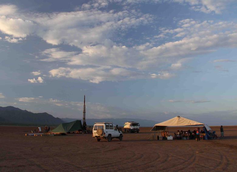 The drill site, Chew Bahar, with tents and vehicles on desert sand. Mountainous peaks appear n the background and the sky s blue, with some light clouds.